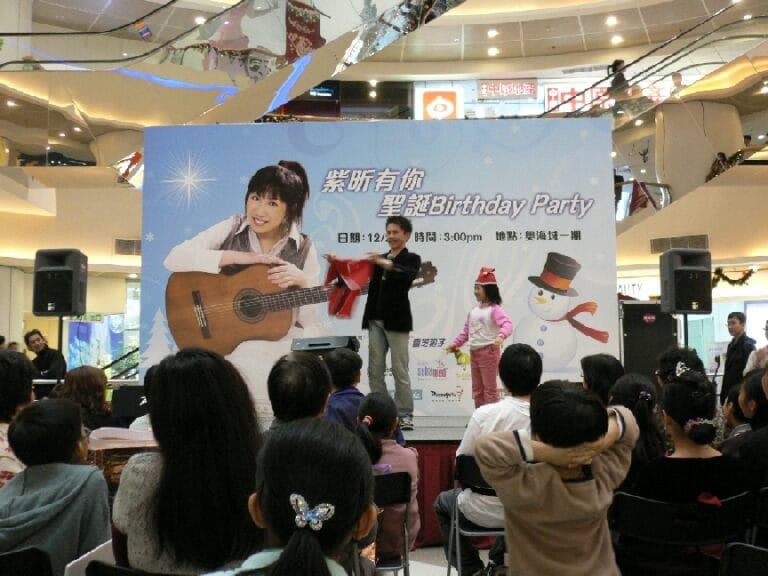 Magician in HK holding a children Santa suit , performing with an audience wearing a red Christmas hat at corporate Christmas birthday party in a shopping mall