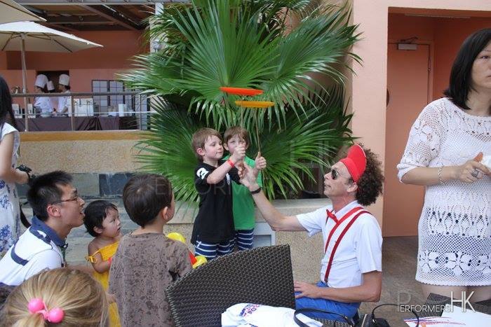 HK roving clown entertainer teaching two kids the Spinning Circus Plate trick at the American Club Independent Day event