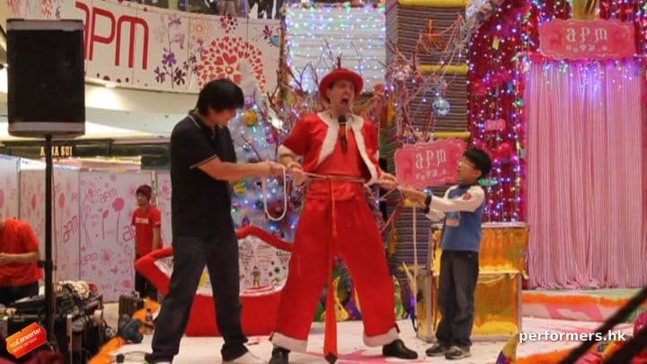 A Hong Kong funny clown performing the rope through the body trick with two volunteer helper from the audience , the try to pull the rope with tight on the entertainers body at a Xmas shopping mall function