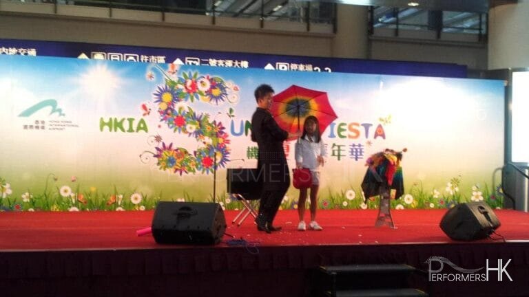 Magician performing magic with an umbrella and an audience holding a red bag standing next to the magic table at a corporate event at Hong Kong Airport Summer Fiesta