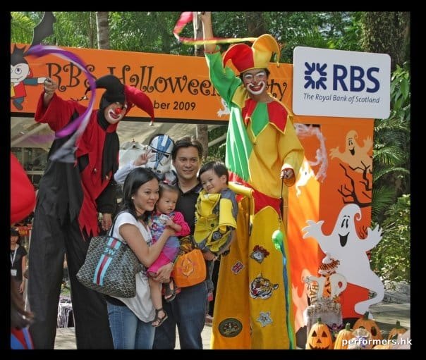 Family of four having a picture taken with two stilt walkers dressed as clowns waving poi