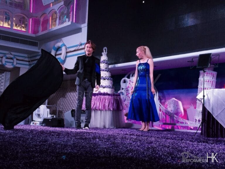 Magician in Hong Kong standing in front of a wedding cake ,holding a black cloth ,performing stage magic with is assistant who wearing a blue dress at a corporate event