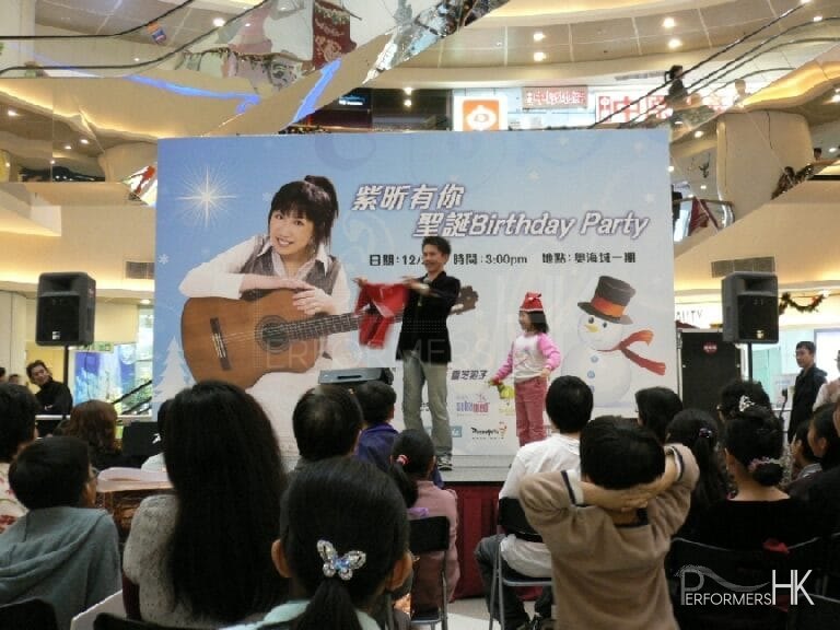 Magician in HK holding a children Santa suit , performing with an audience wearing a red Christmas hat at corporate Christmas birthday party in a shopping mall