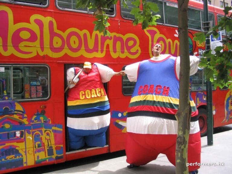Hong Kong stilt-walker in a blue inflatable coach costume pulling other stilt-walker in a red inflatable coach costume from a bus