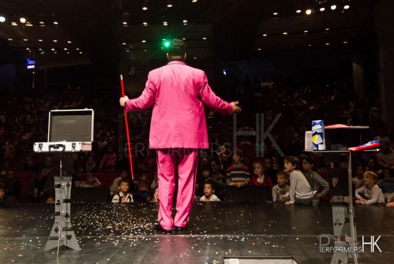 Children looking at the magician in Hong Kong who holding a red wand , standing next to the magic case , performing at the corporate kids event